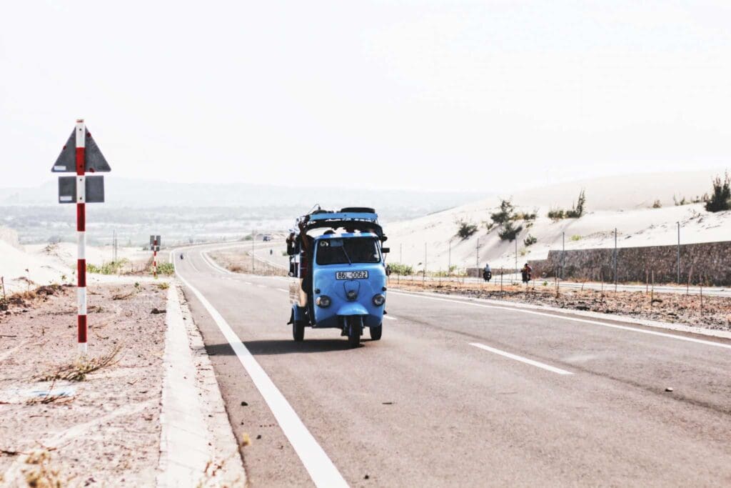 A small blue three-wheeled vehicle travels alone on a deserted, winding road through a sandy desert landscape. Sparse shrubs and road signs are visible along the roadside under a clear sky.