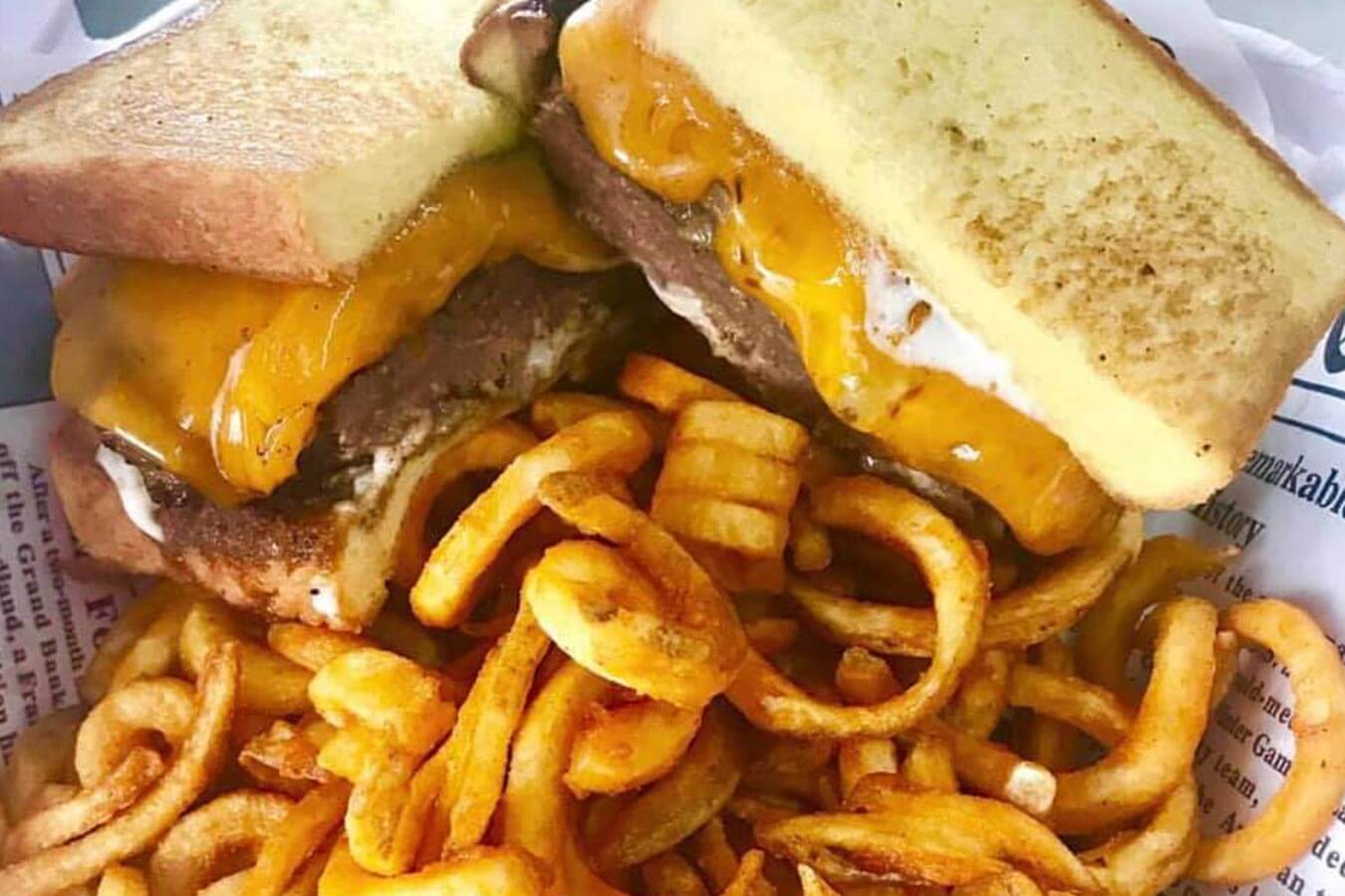 Curly fries piled in front of two grilled cheese sandwiches stuffed with burger patties and melted cheddar cheese, resting on a piece of paper.