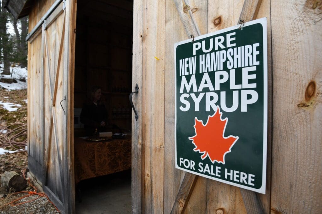 A wooden shed with an open door displays a sign reading Pure New Hampshire Maple Syrup for Sale Here with a red maple leaf. Inside, a person stands behind a table. Snow is visible on the ground outside.
