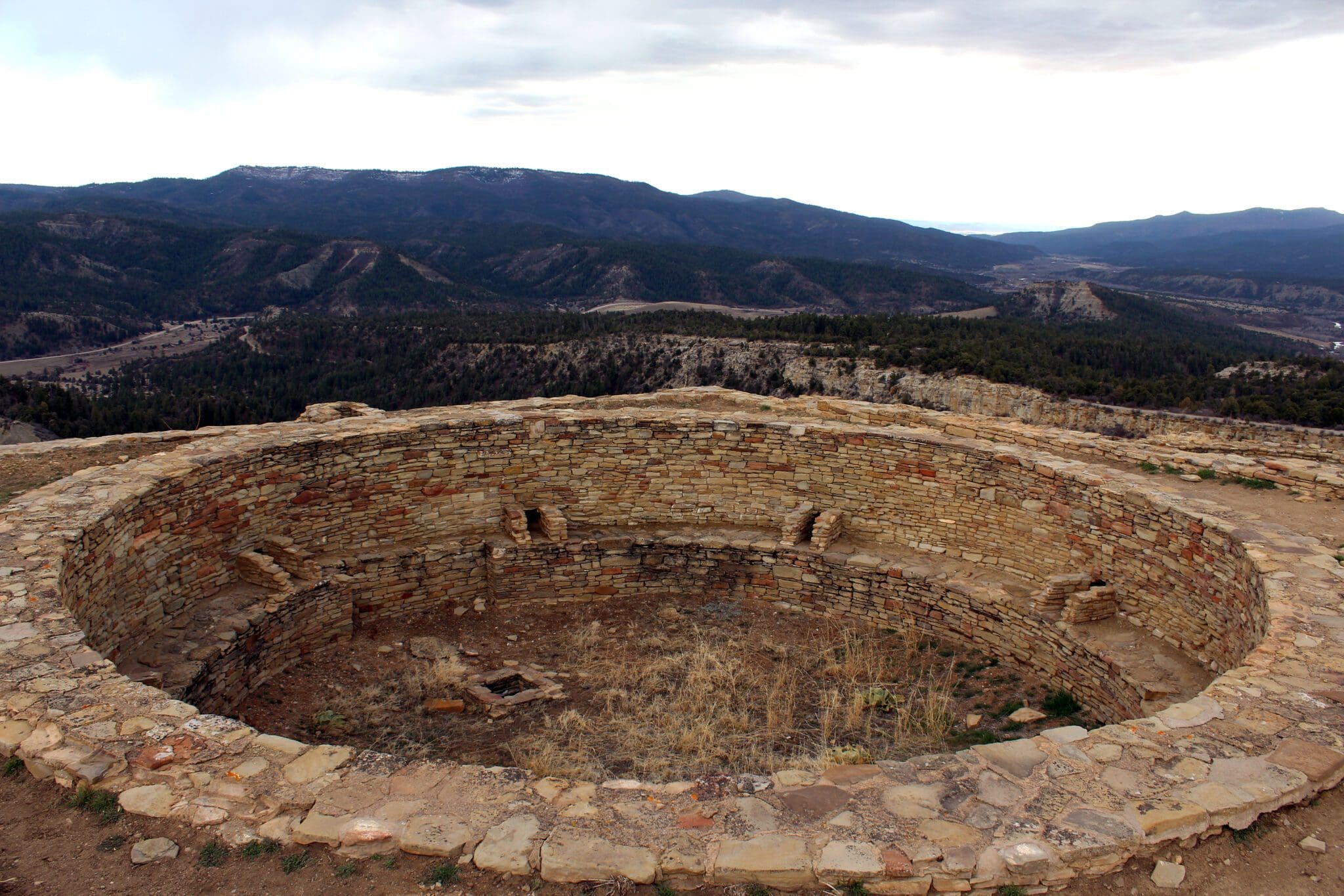 A circular ancient stone structure with a sunken interior, set on a hilltop. Surrounding it are expansive views of rolling hills and distant mountains under a cloudy sky. Sparse vegetation is visible around the structure.