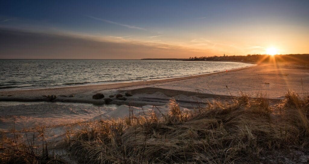 Sunset over a tranquil beach with gentle waves meeting the shore. In the foreground, tall grasses atop sand dunes add texture. The sky transitions from soft blue to warm orange as the sun sets on the horizon.