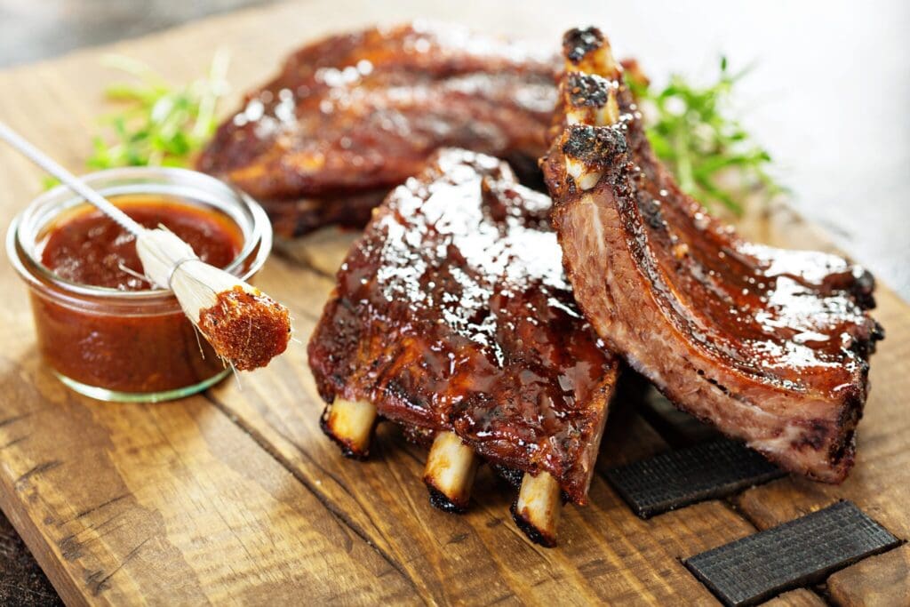 Grilled ribs with a glossy barbecue sauce on a wooden board, garnished with fresh herbs. A small brush dipped in barbecue sauce rests near a glass bowl filled with the sauce.