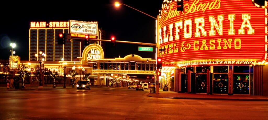 Street scene at night with bright neon lights of John Boyds California Hotel & Casino and Main Street Station in Las Vegas. Illuminated signs and a few cars are visible on the street, surrounded by buildings with bright marquee lights.