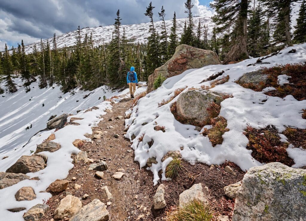 A person in a blue jacket and backpack hikes along a snow-dusted trail surrounded by evergreen trees and large rocks, with a mountain visible in the background under a cloudy sky.