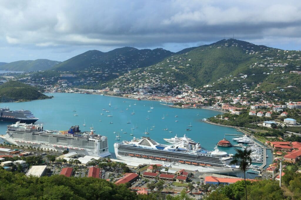 Aerial view of a tropical harbor with multiple cruise ships docked and various smaller boats in the water. The harbor is surrounded by lush green hills and a town with numerous buildings. The sky is partly cloudy.