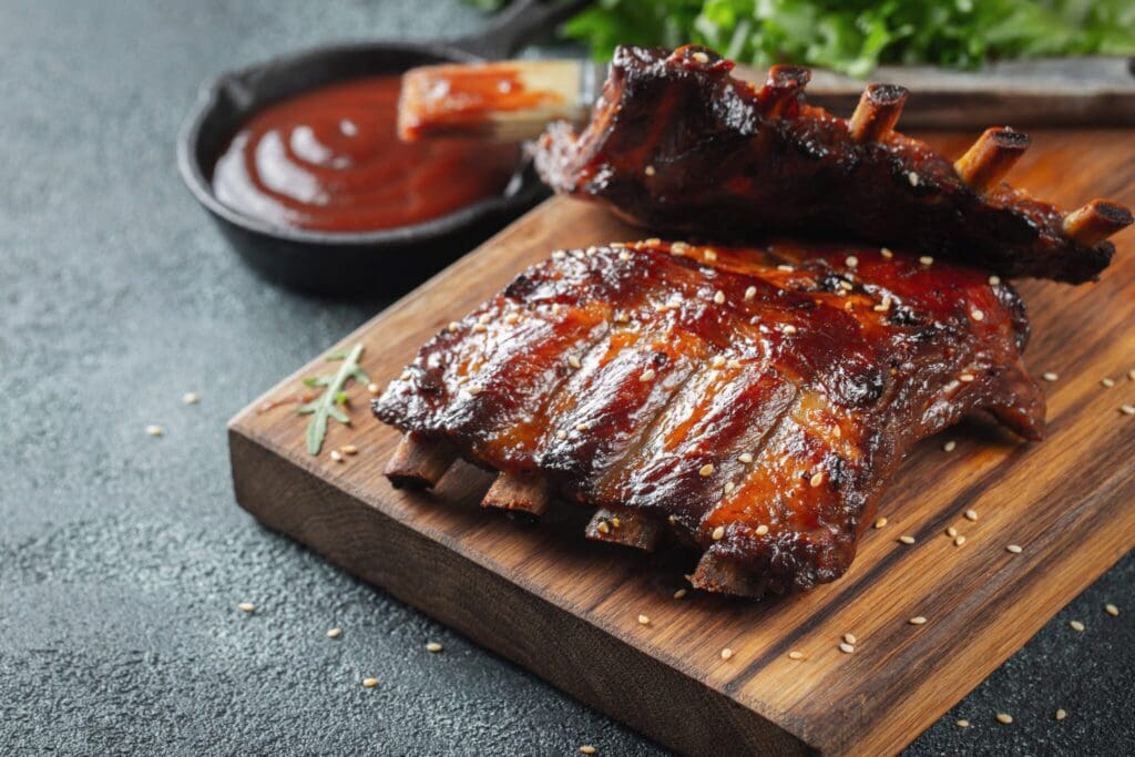Grilled barbecue ribs glazed with sauce sit on a wooden board, garnished with sesame seeds. In the background, there’s a bowl of barbecue sauce with a brush and some fresh leafy greens.