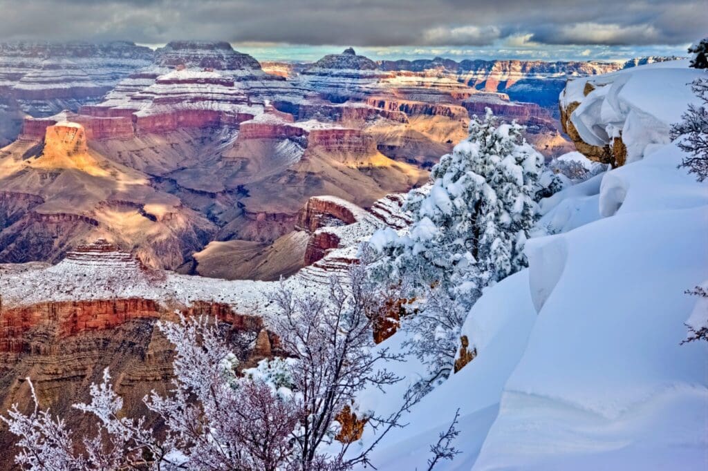 Snow blankets the rocky cliffs and bushes of the Grand Canyon under a cloudy sky. The light casts shadows, highlighting the layers of colorful rock formations and snow-covered terrain, creating a striking contrast between the icy foreground and distant canyon.