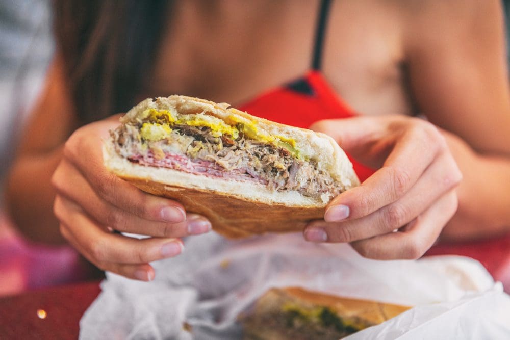 A person in a red top holding a partially eaten sandwich filled with shredded meat, sliced ham, and mustard. The sandwich is on white wrapping paper, and the background is blurred.