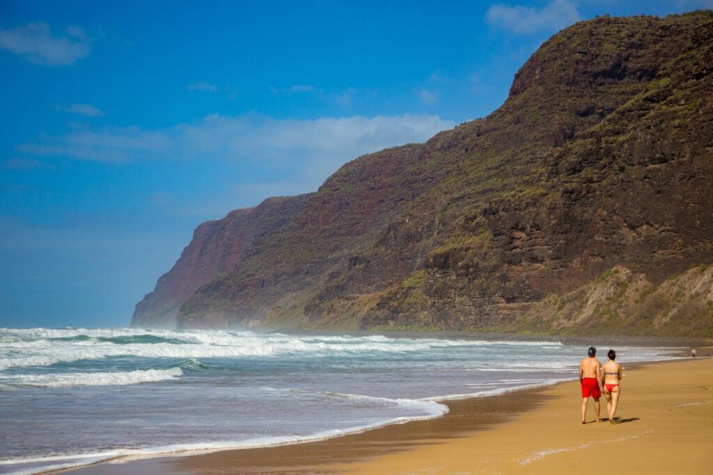 Two people walk along a sandy beach with gentle waves on the left. Cliffs rise steeply on the right, covered in greenery. The sky is blue with a few clouds.