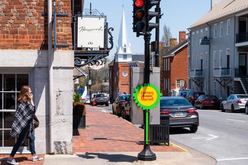 A woman walks on a sidewalk, passing a brick building with a Haysworths sign. Across the street, several parked cars are visible. A Farmers Market sign is attached to a pole. In the background, a church steeple and more buildings are seen.