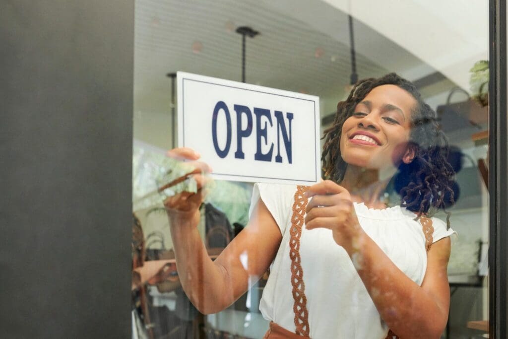 Woman with curly hair smiles while placing an OPEN sign on the inside of a glass door, suggesting a shop or business is ready for customers. Shes wearing a white top with decorative straps.