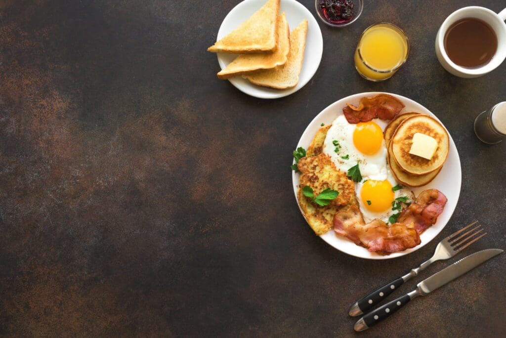 A breakfast plate with pancakes topped with butter, two sunny-side-up eggs, bacon, and toast. Accompanied by drinks, including orange juice and coffee, with utensils placed beside the plate on a dark surface.