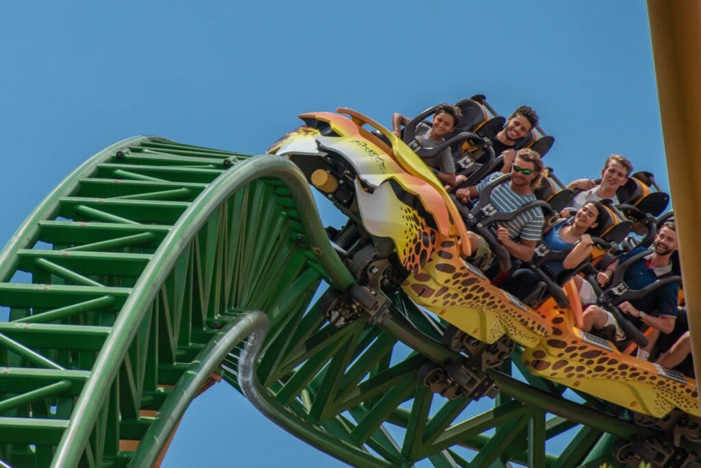 People are riding a roller coaster with a vibrant design resembling a yellow and green cheetah. The roller coaster is ascending a green loop against a clear blue sky. The riders look excited and are seated in a row.