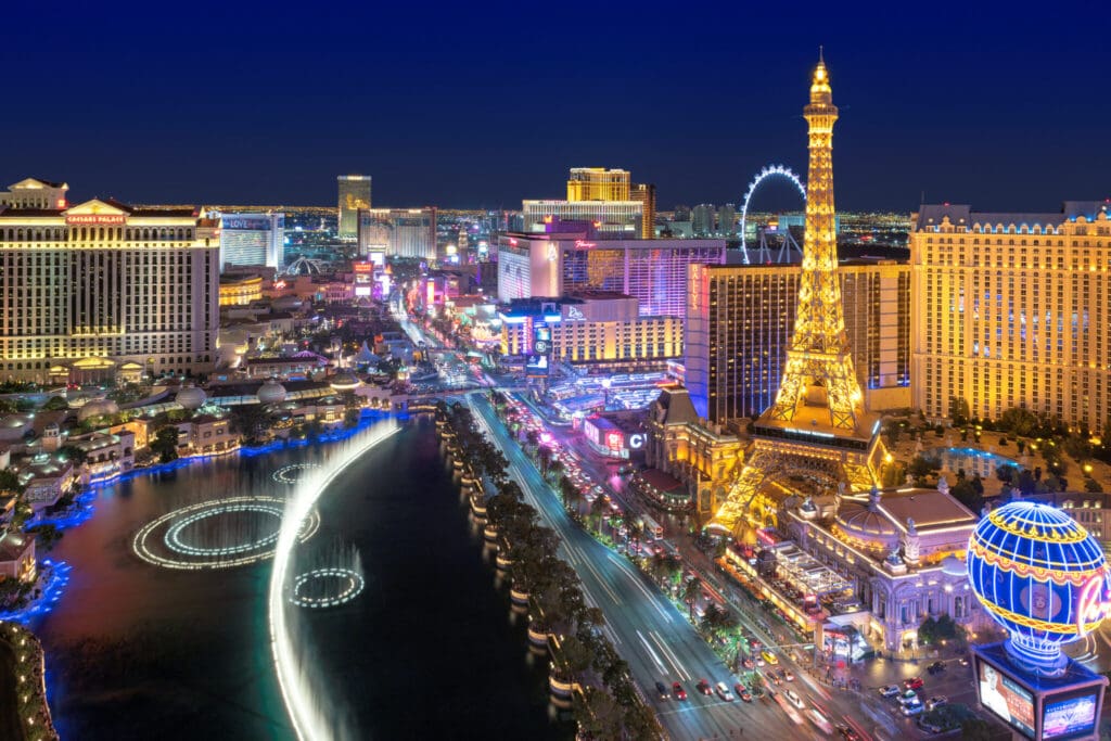 Aerial view of the Las Vegas Strip at night, featuring brightly lit casinos and hotels, including replicas of the Eiffel Tower and the Arc de Triomphe. The High Roller Ferris wheel and water fountains are also visible in the vibrant scene.