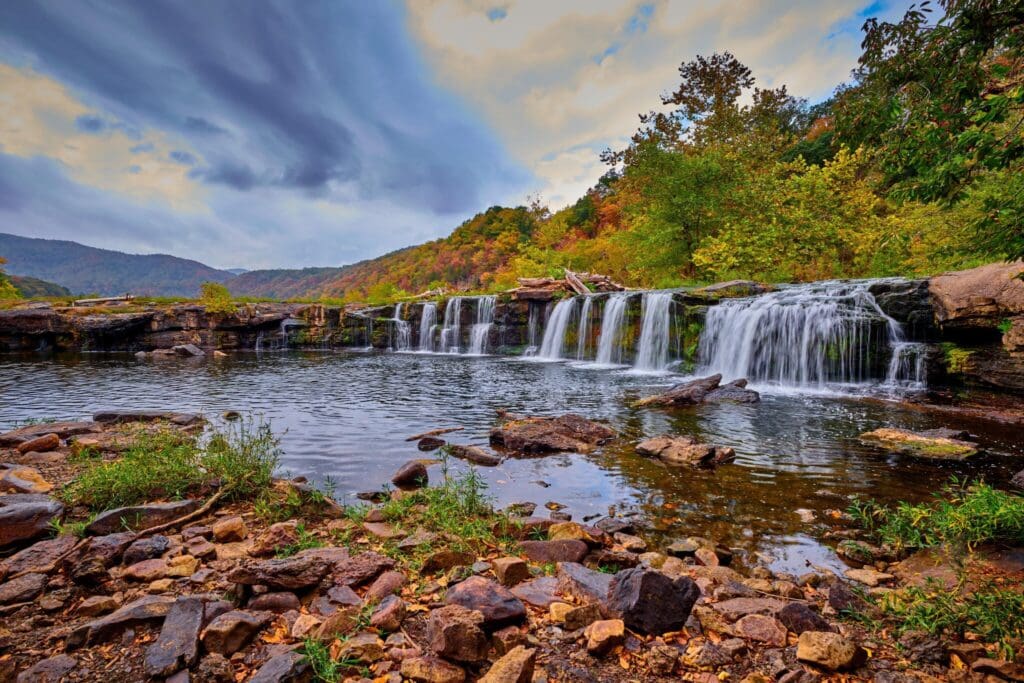A serene landscape featuring a cascading waterfall flowing over rocks into a calm pool, surrounded by lush greenery and colorful autumn foliage. A cloudy sky looms overhead, enhancing the tranquil and natural ambiance of the scene.