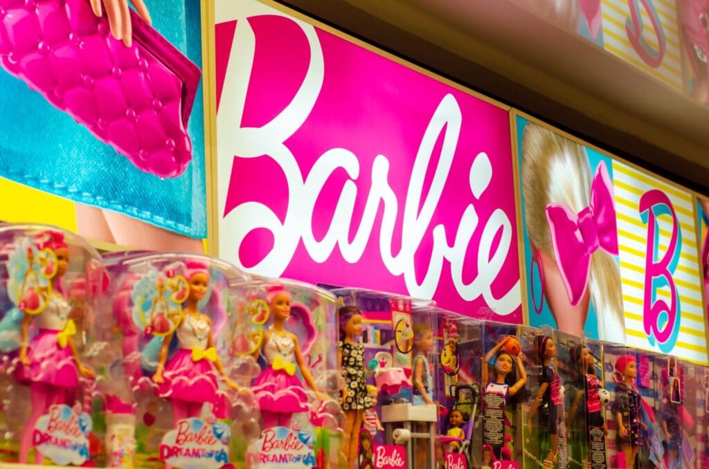 A store display featuring various Barbie dolls in colorful packaging. Above the dolls, there is a prominent pink and white sign with Barbie written on it, alongside images of a purse and a pink bow.
