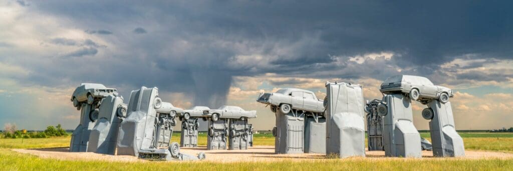 A panoramic view of Carhenge, an art installation featuring vintage cars stacked and arranged in a stonehenge-like formation on a grassy field. Dark storm clouds gather in the sky, creating a dramatic backdrop.