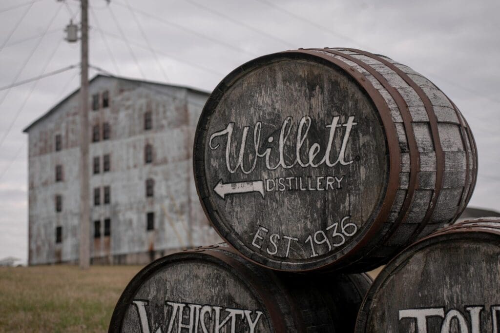 Three whiskey barrels with Willett Distillery EST 1936 written on them are in the foreground. An old, weathered industrial building stands in the background under a cloudy sky.