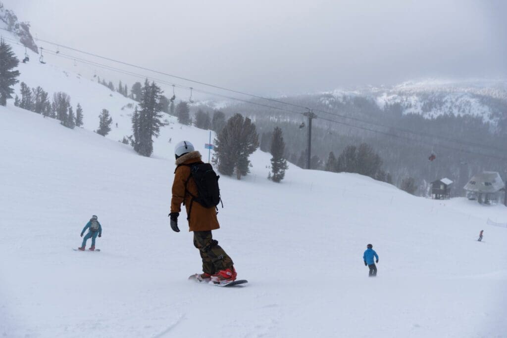 A person in a brown jacket and camouflage pants snowboards down a snowy mountain slope. In the background, two other people in blue jackets are skiing. The sky is overcast, with trees and a mountain visible.