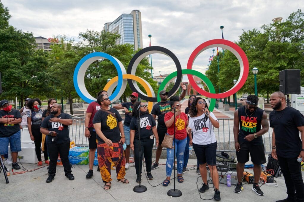 A diverse group of people stand singing and speaking into microphones in front of large Olympic rings outside. They wear shirts with various slogans, including Black Lives Matter. Trees and a building are visible in the background.