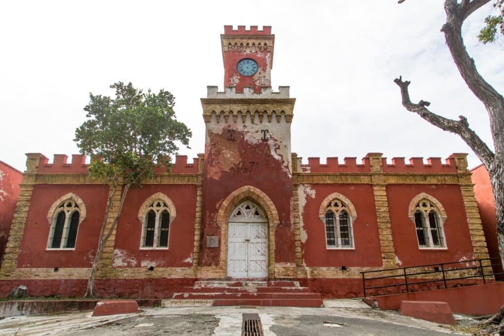 The image shows a red brick building with a tall clock tower and gothic-style arched windows. The facade is weathered, with some peeling paint. A bare tree stands to the right. The year 1871 is visible above the main entrance.