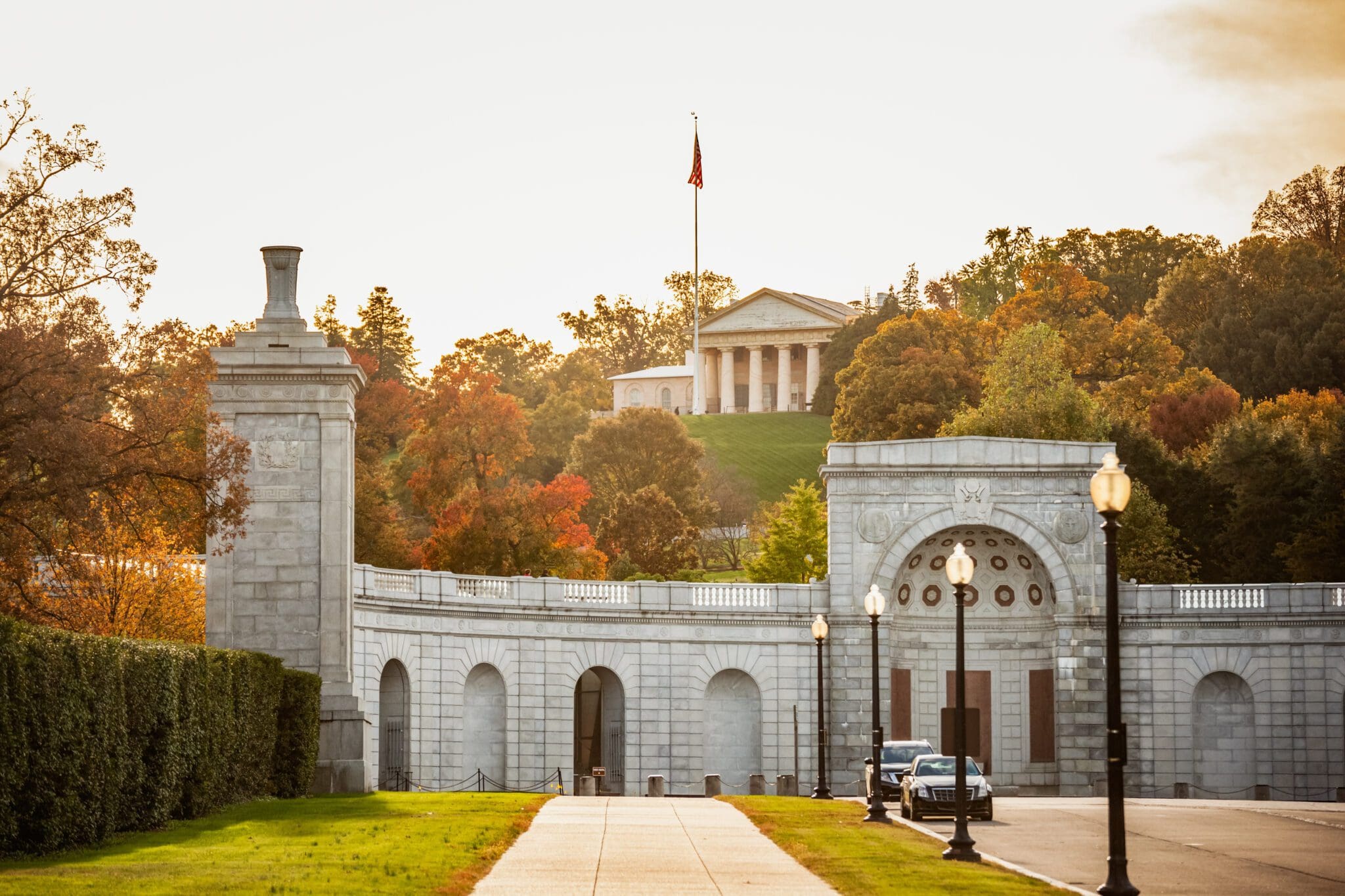 A sunny view of a large white stone entryway with arches and columns, leading to a hilltop building resembling a Greek temple with columns. The scene is framed by colorful autumn trees. A few cars are parked near the entrance.