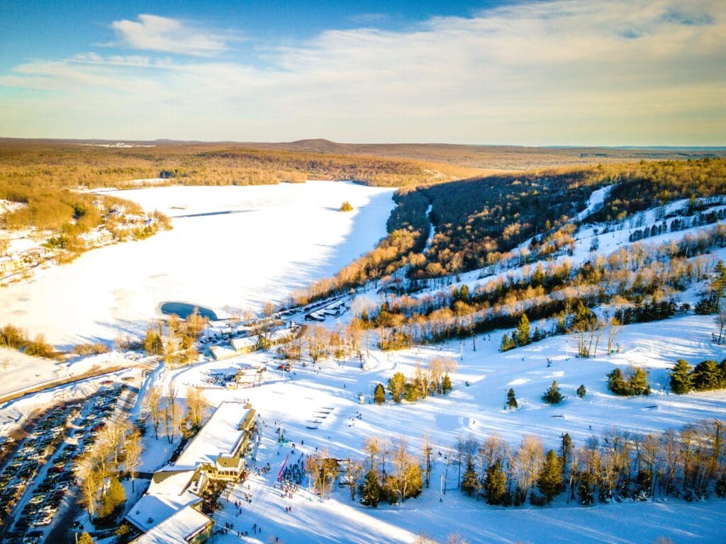 Aerial view of a snowy landscape featuring a frozen lake surrounded by wooded hills under a clear blue sky. In the foreground, ski slopes and buildings are visible, with parking areas and people engaging in winter activities.