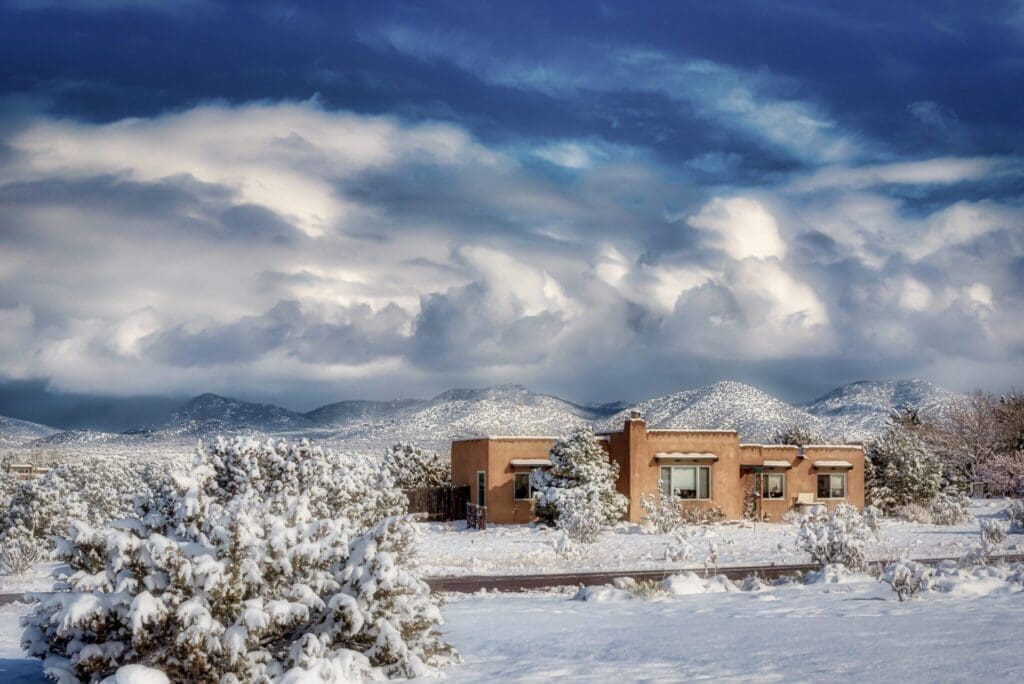 Adobe-style house in a snow-covered landscape with sparse trees. Mountains are visible in the background under a dramatic, cloudy sky.