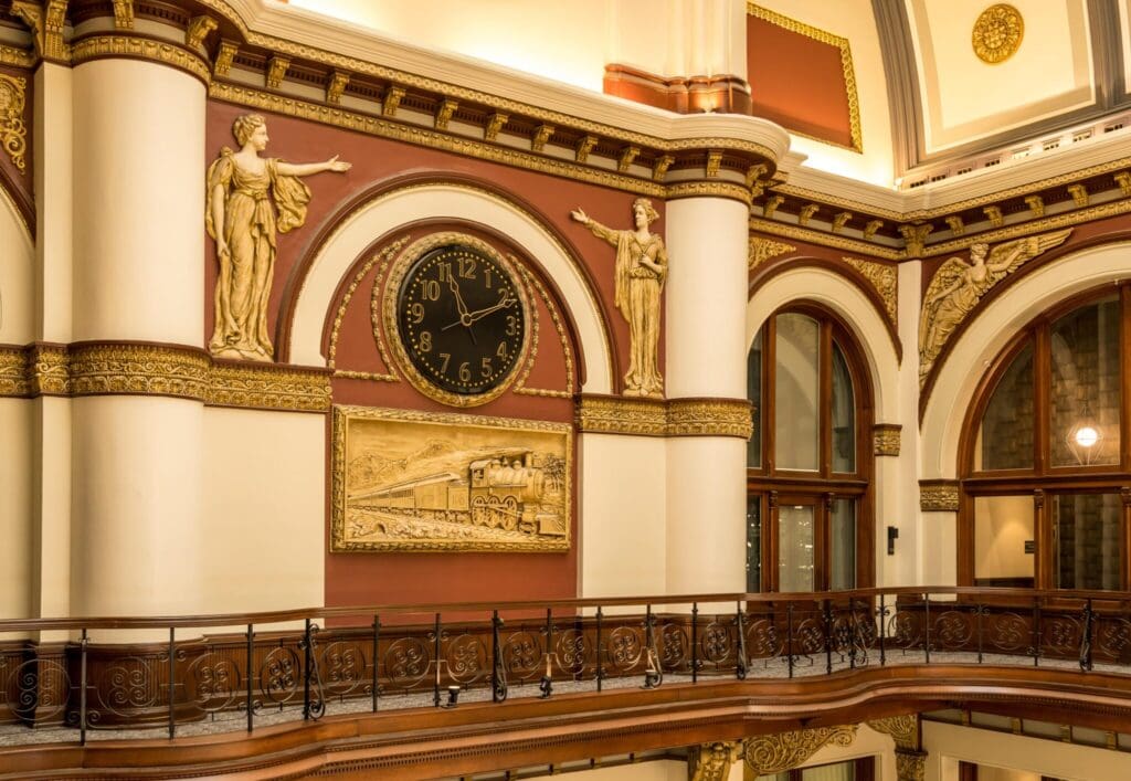 Interior of an ornate building with a large round clock, gold statues on either side, a detailed golden train relief below the clock, and arched windows. The decor is lavish with white columns and decorative molding.