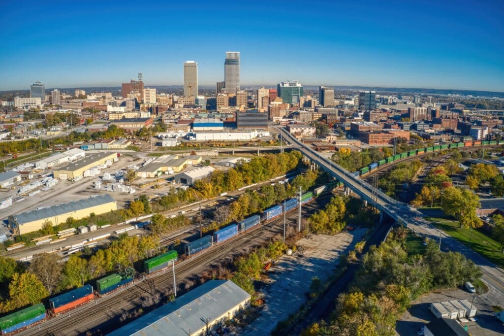 Aerial view of a cityscape with a mix of skyscrapers and low-rise buildings under a clear blue sky. A long bridge runs over a railway yard with colorful freight containers. Greenery lines parts of the rail area.