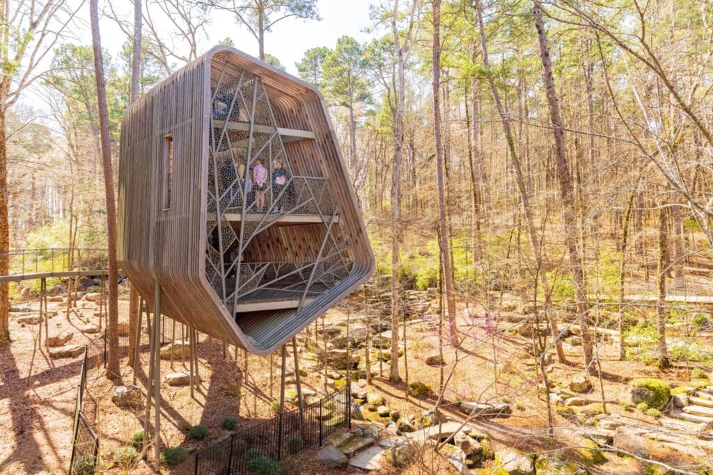 A modern, geometric treehouse sits elevated among tall trees in a woodland setting. Its design features large, angular windows with a mesh-like exterior, blending seamlessly with the natural surroundings. People can be seen inside the structure.