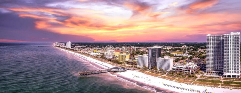 Aerial view of a coastline at sunset, featuring a vibrant orange and pink sky. The shoreline is dotted with tall buildings and a pier extending into the ocean. The beach is lined with white sand and beachgoers. Urban skyline fades into greenery.