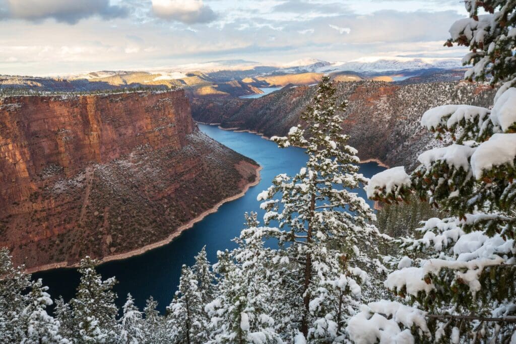 Snow-covered trees frame a scenic view of a winding river below steep red cliffs. The landscape stretches to distant mountains under a cloudy sky, with patches of sunlight highlighting the terrain.
