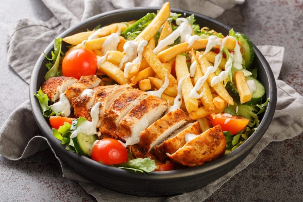 A bowl filled with sliced grilled chicken, crispy fries, cherry tomatoes, and fresh greens, all drizzled with a creamy dressing, rests on a gray cloth surface.