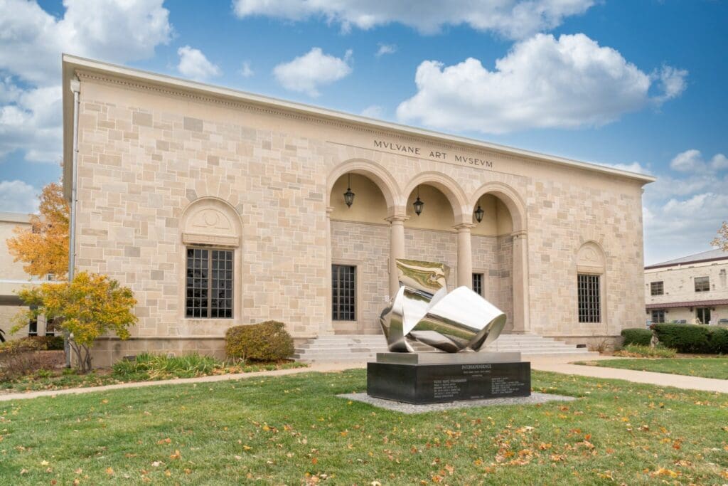 The image shows the Mulvane Art Museum, a classic stone building with arched windows and an entrance. A modern metallic sculpture stands on the lawn in front. The sky is partly cloudy, with fallen leaves scattered on the grass.