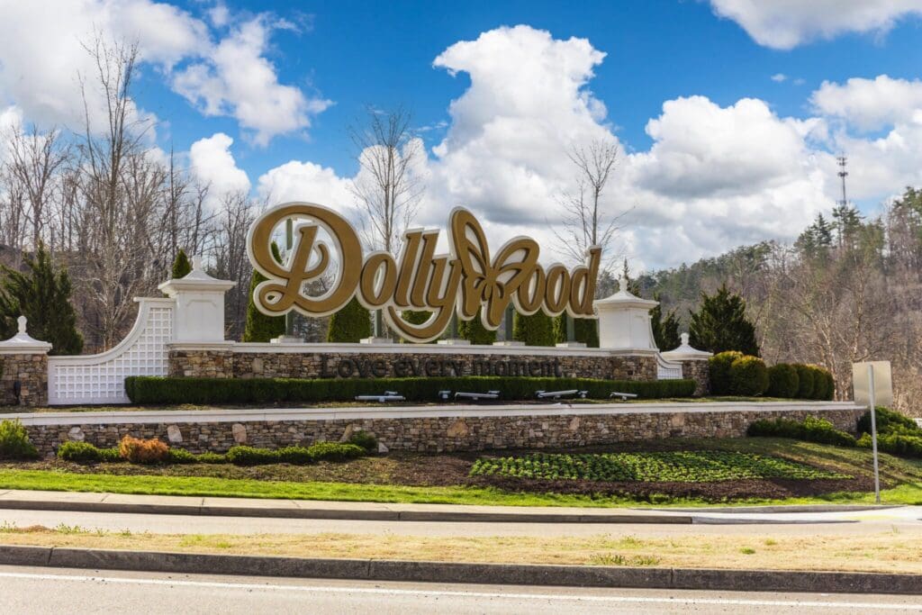 The image shows the entrance sign to Dollywood, a theme park. The large golden letters of Dollywood stand out, with Love every moment below. The sign is surrounded by landscaped greenery, with a cloudy sky in the background.