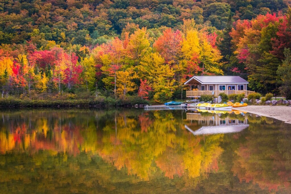 A wooden cabin by a lake is surrounded by vibrant autumn trees in shades of red, orange, and yellow. The calm water reflects the colorful foliage and the cabin. Several kayaks are lined up on the sandy shore.