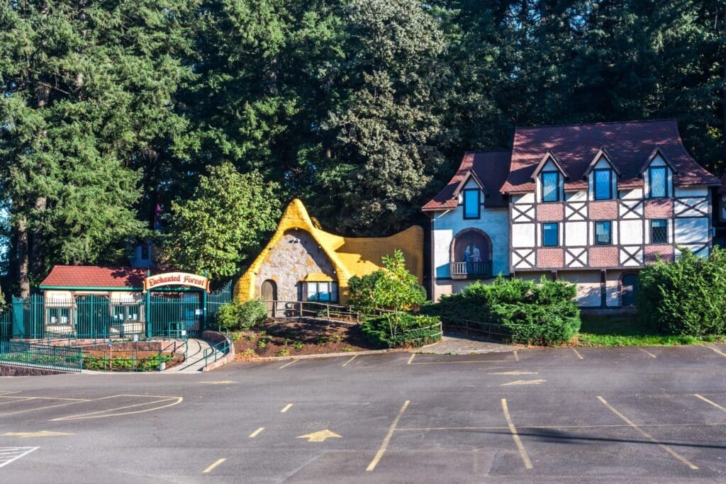 A quaint village-like scene featuring a Tudor-style building with a red roof and a whimsical, yellow-roofed cottage. Dense trees form the backdrop, and a sign reads Enchanted Forest near a gated entryway. A large empty parking lot is in the foreground.