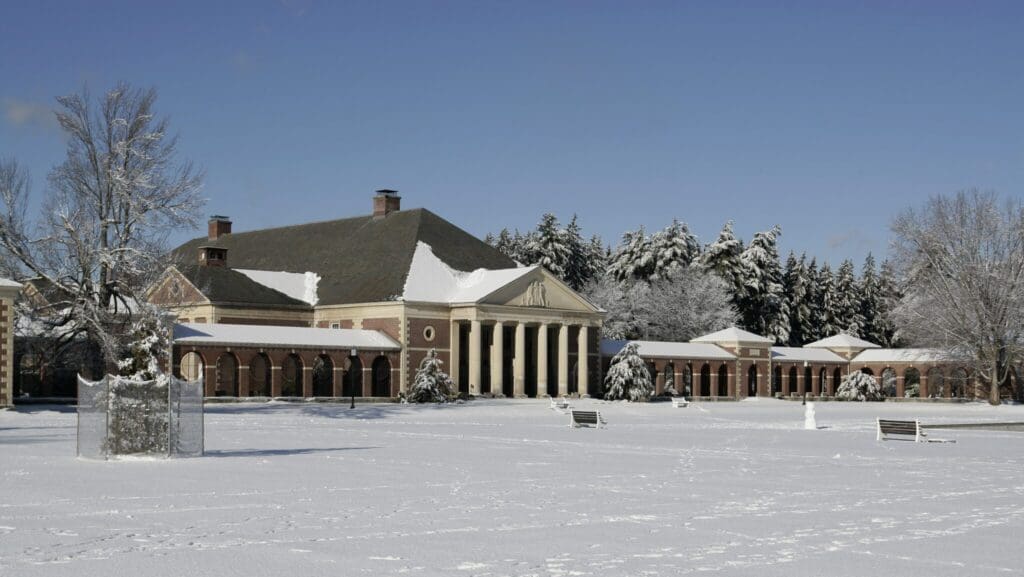 A large, stately building with columns is surrounded by snow-covered trees and grassy areas. Benches and a snow-dusted path are visible in the foreground under a clear blue sky.