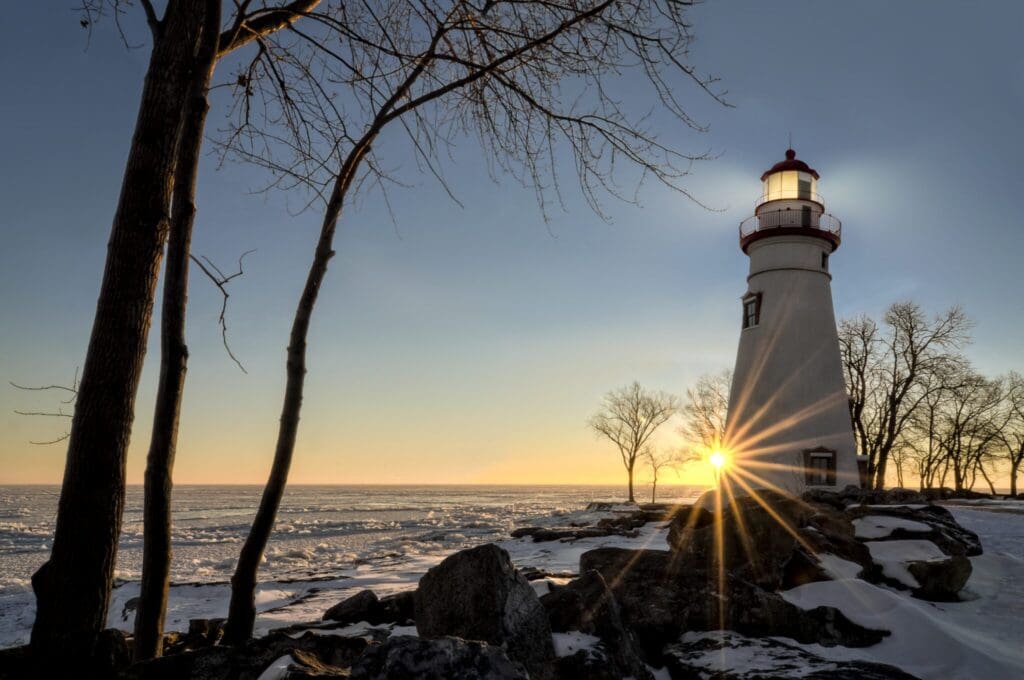 A lighthouse stands by a frozen, rocky shoreline at sunset. Bare trees surround it, and the sun peeks through, casting a warm glow and starburst effect. The sky is clear, transitioning from blue to orange near the horizon.