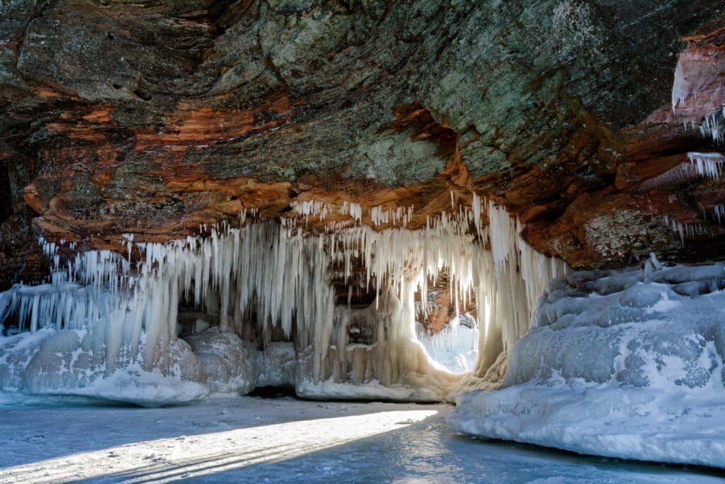 Icicles hang from the red and green rocky ceiling of an ice cave, with sunlight streaming through a small opening. The ground is frozen, and the ice formations create a natural, sculpted look.