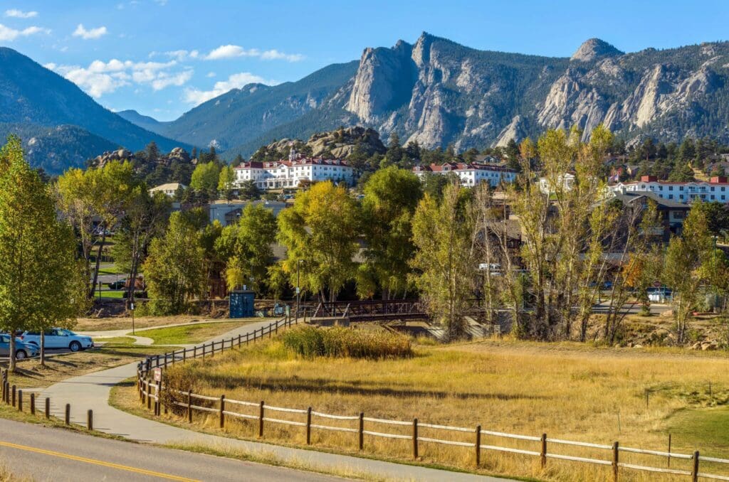 A scenic view featuring a path bordered by grass and wooden fences leading to a small bridge. In the background, a cluster of buildings is set against a backdrop of rugged, tree-covered mountains under a clear blue sky.