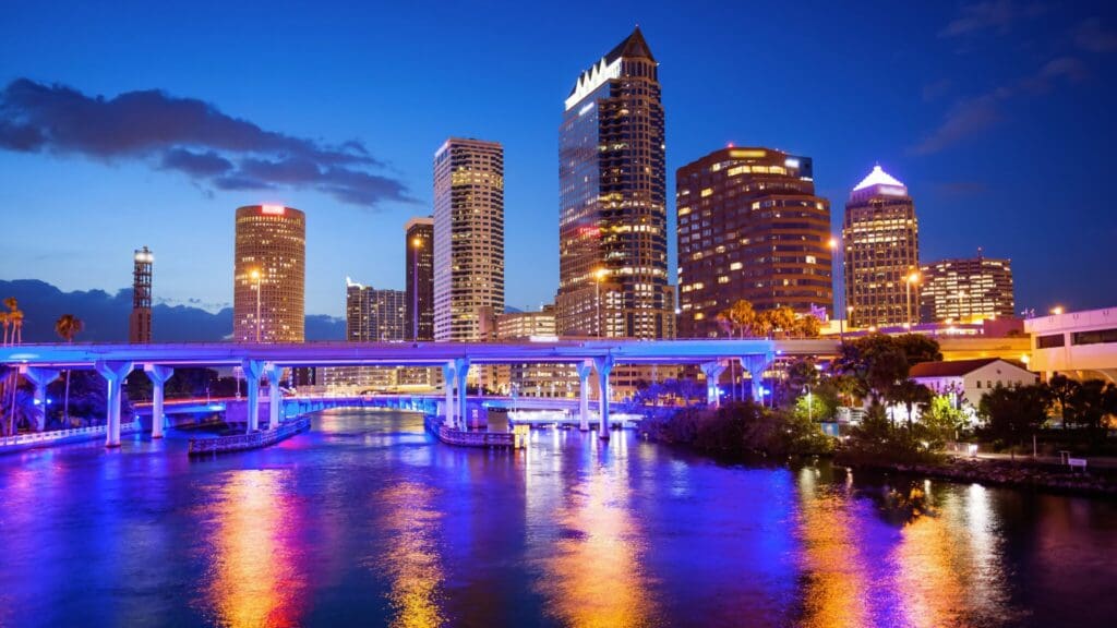 City skyline at dusk with illuminated skyscrapers reflected in a river. A bridge spans the water, with lights casting blue and purple hues. The sky transitions from deep blue to orange on the horizon.