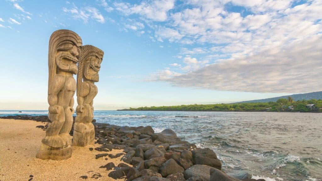Two wooden tiki statues stand on a sandy beach with black rocks, overlooking the ocean. The sky is partly cloudy, and green vegetation lines the distant shore.