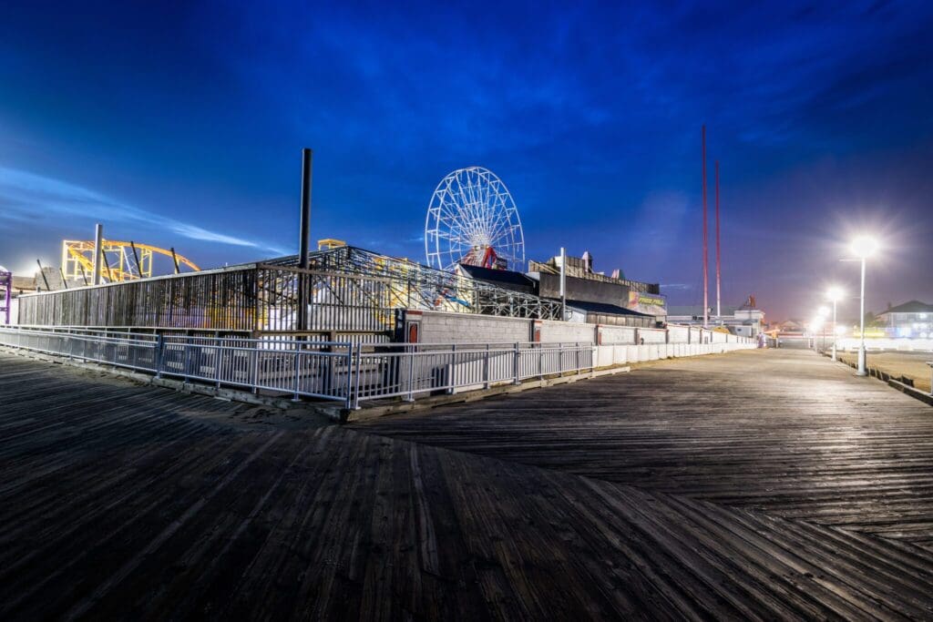 Night view of a boardwalk leading to an amusement park with a Ferris wheel and roller coaster. The sky is dark blue, and lamp posts illuminate the scene. The area appears empty and serene.