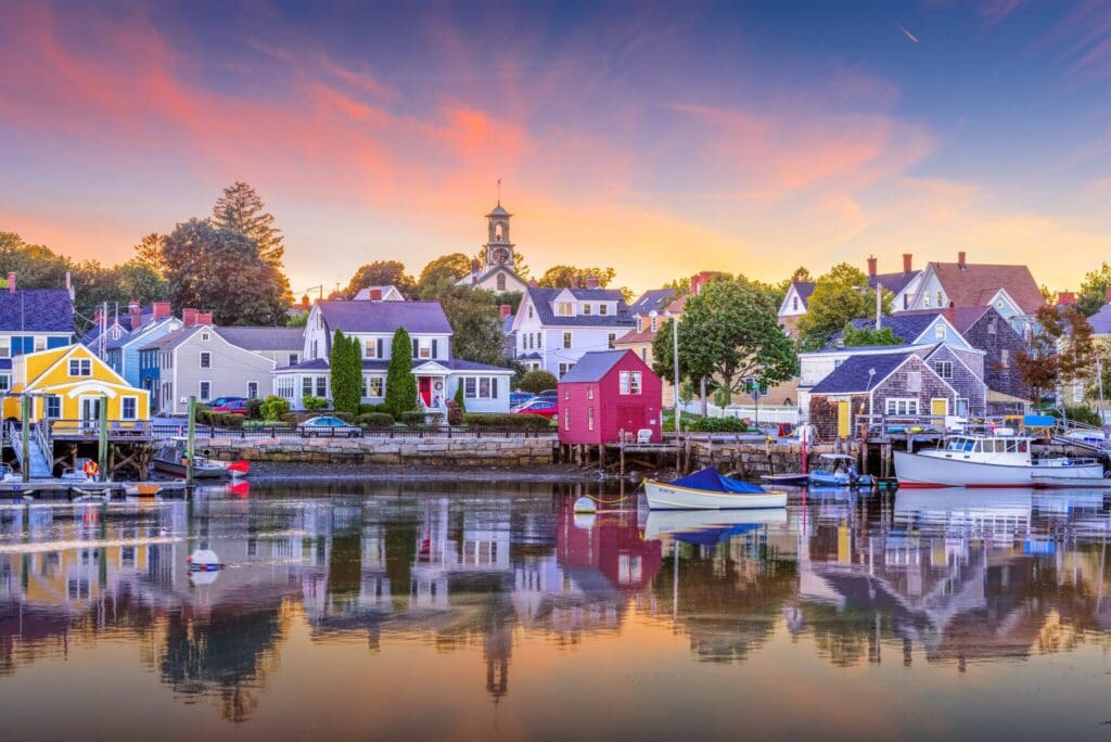 A picturesque harbor at sunset features quaint, colorful houses and boats reflected in the calm water. The sky is a gradient of orange to blue with wispy clouds, and a small church steeple is visible among the houses.