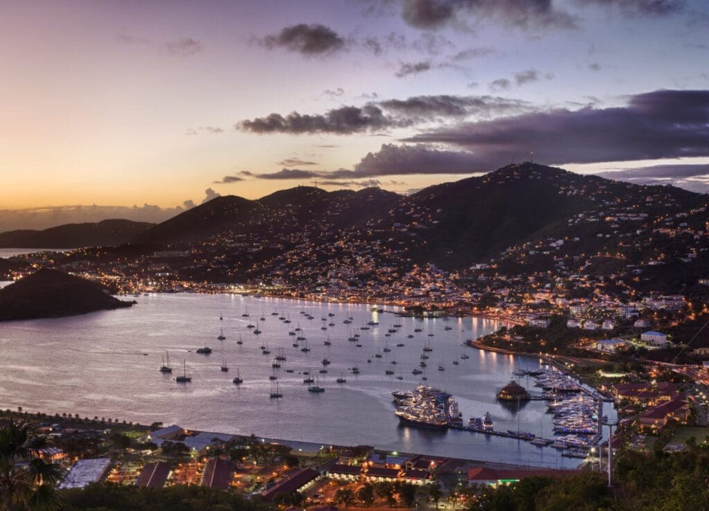 Aerial view of a picturesque coastal town at dusk, with a bay filled with boats. Hills surrounding the town are adorned with scattered lights. The sky transitions from golden hues to deep purple, reflecting on the calm water.