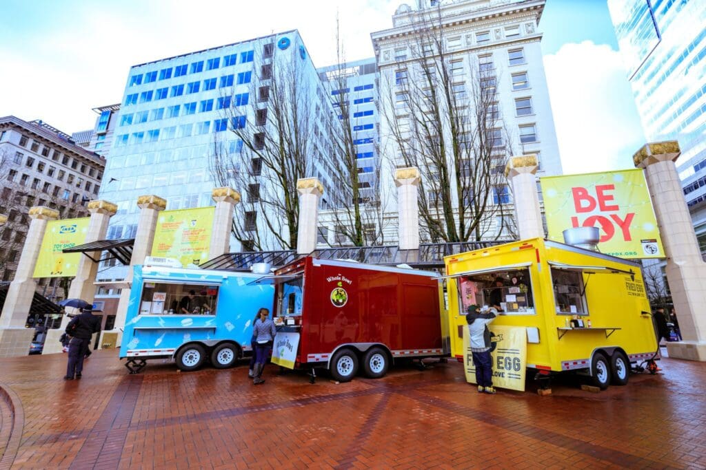 Food trucks in a city square with tall buildings and trees in the background. Three trucks in blue, red, and yellow serve customers. People with umbrellas stand around, and banners with Be Joy are visible on the building facades.