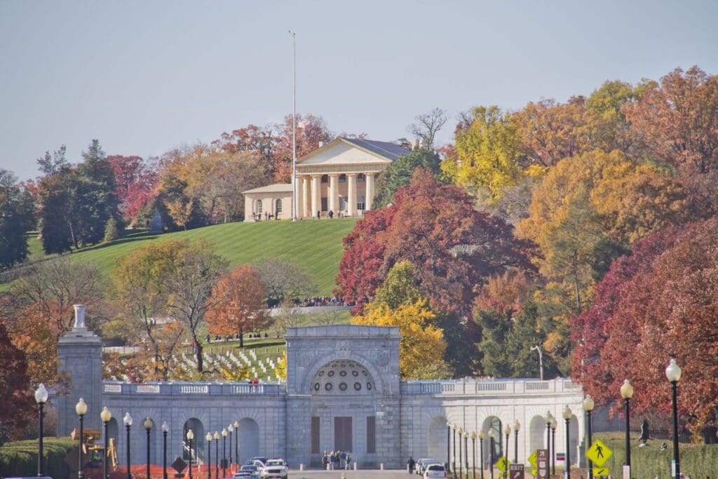 A grand neoclassical building with columns is situated atop a hill, surrounded by vibrant autumn trees in shades of red, orange, and yellow. In the foreground, an ornate stone gate with arches stands amidst a landscaped area.