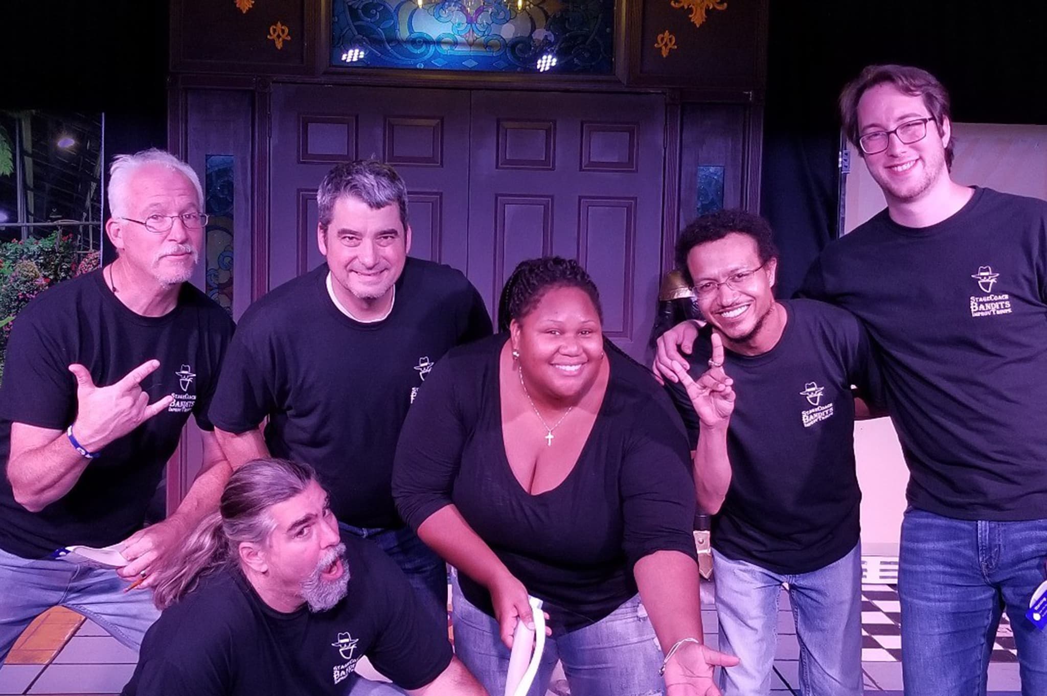 Six people wearing similar black shirts pose and smile on a stage with a decorative backdrop. They appear to be a diverse group of men and a woman, some making playful gestures towards the camera.